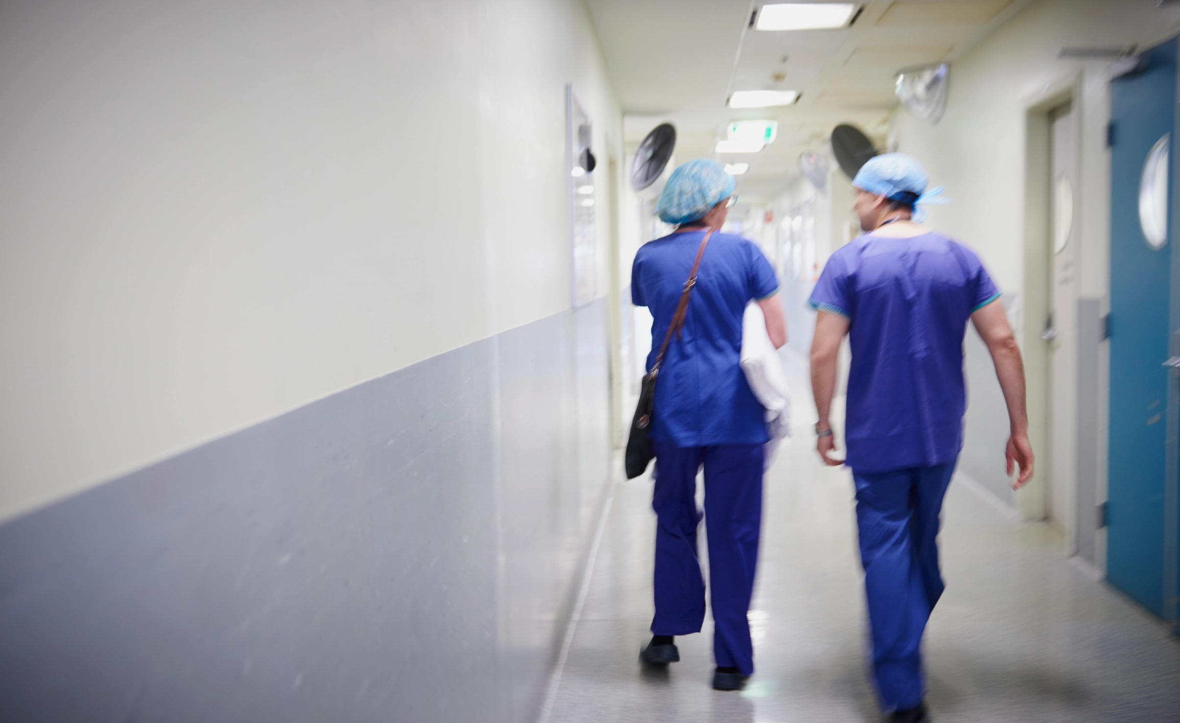 a male and female doctor in scrubs walk down a hospital corridor, away from the camera