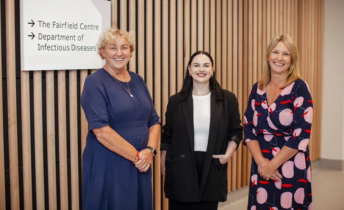 Three professionally dressed trial coordinators stand together, smiling, in front of a sign for "The Fairfield Centre Department of Infectious Diseases," against a modern wooden panel backdrop.