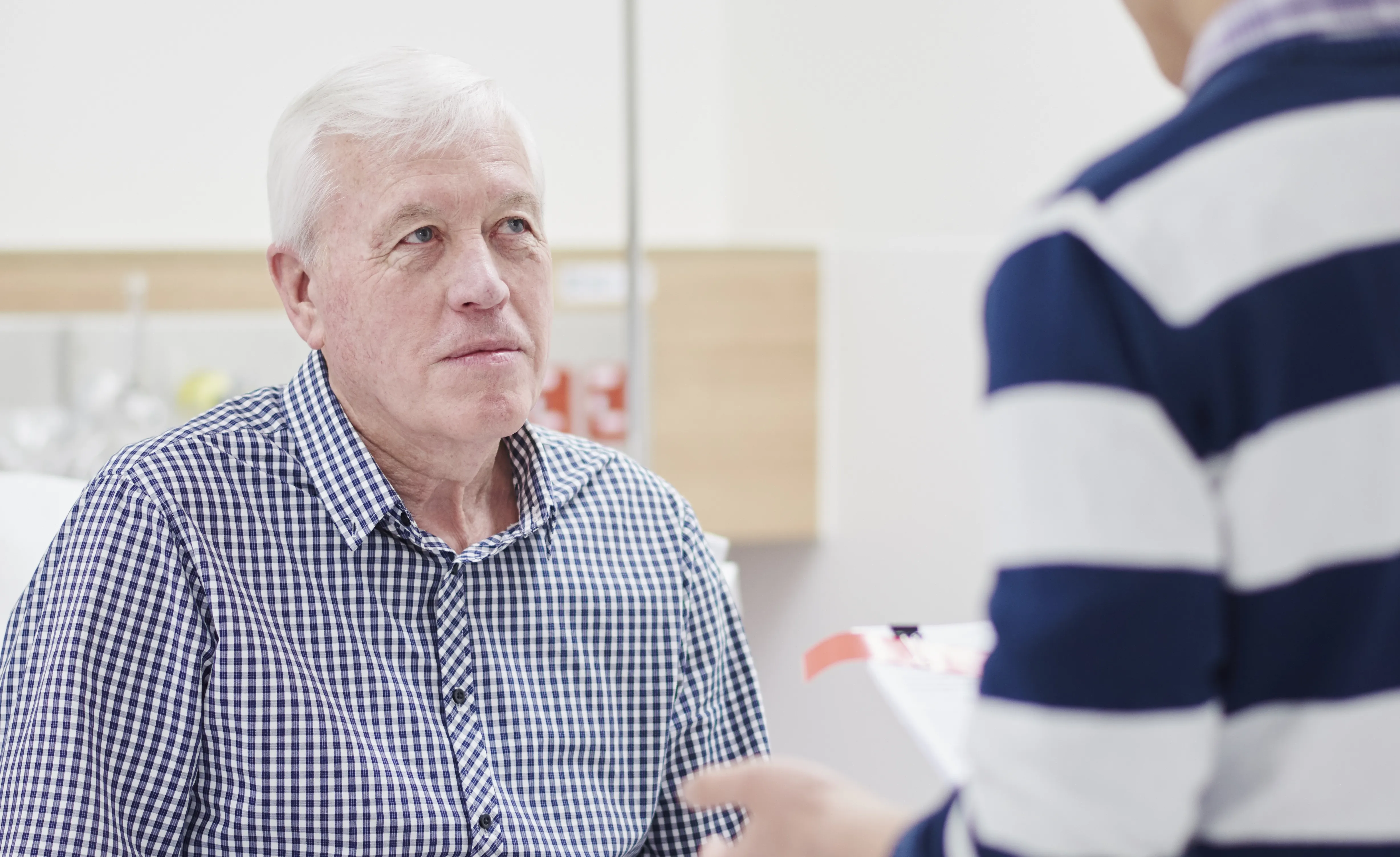 An older patient in a checkered shirt listens attentively to a discussion with an anaesthetist holding a document in a clinical setting.