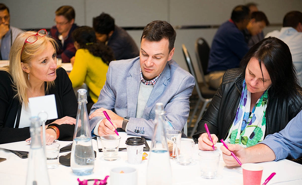 A group of professionals at a round table, engaged in discussion and note-taking at a table.