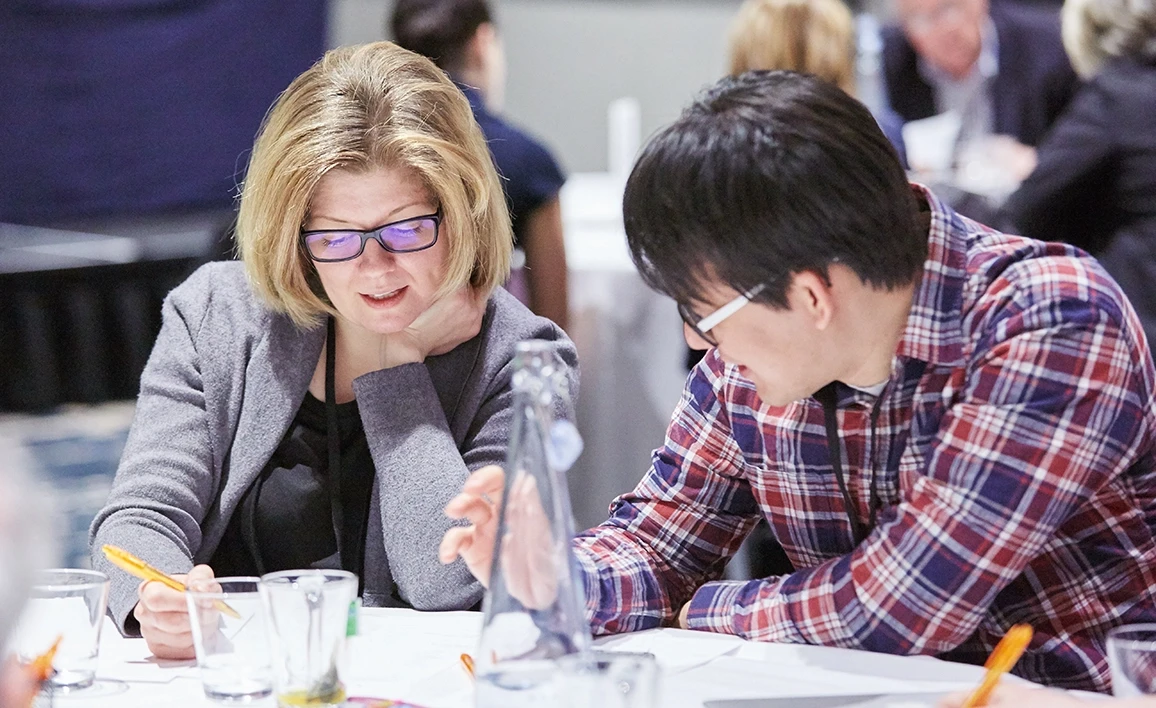 Two professionals collaborate at a table, reviewing documents and taking notes in a conference or workshop setting, with other attendees in the background.