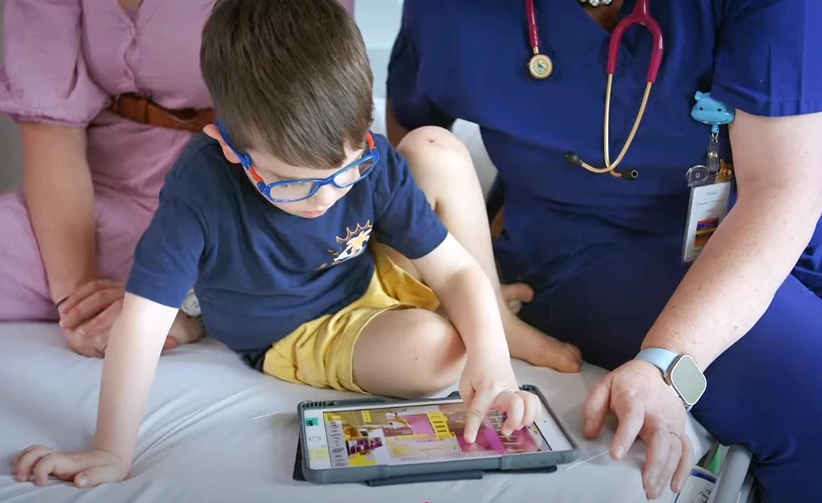 A young child wearing glasses interacts with a tablet while sitting on a hospital bed, accompanied by a caregiver and an anaesthetist in scrubs with a stethoscope.