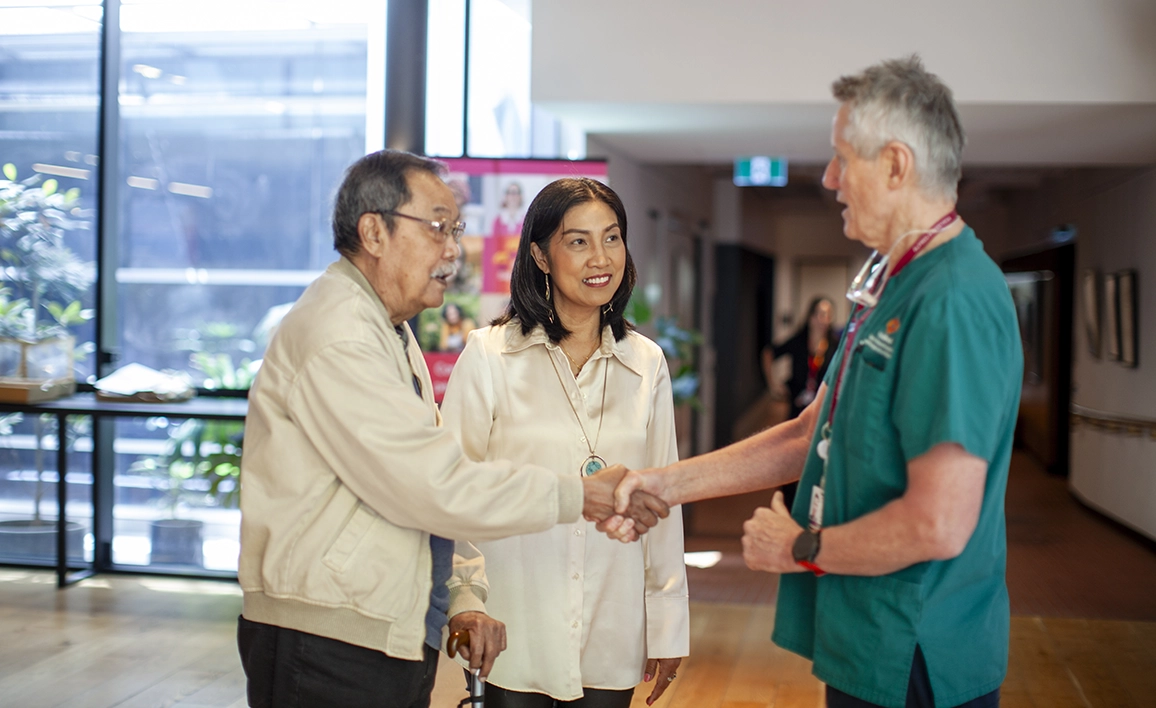 A friendly interaction in a medical setting, where an older man shakes hands with an anaesthetist while a caregiver smiles beside them