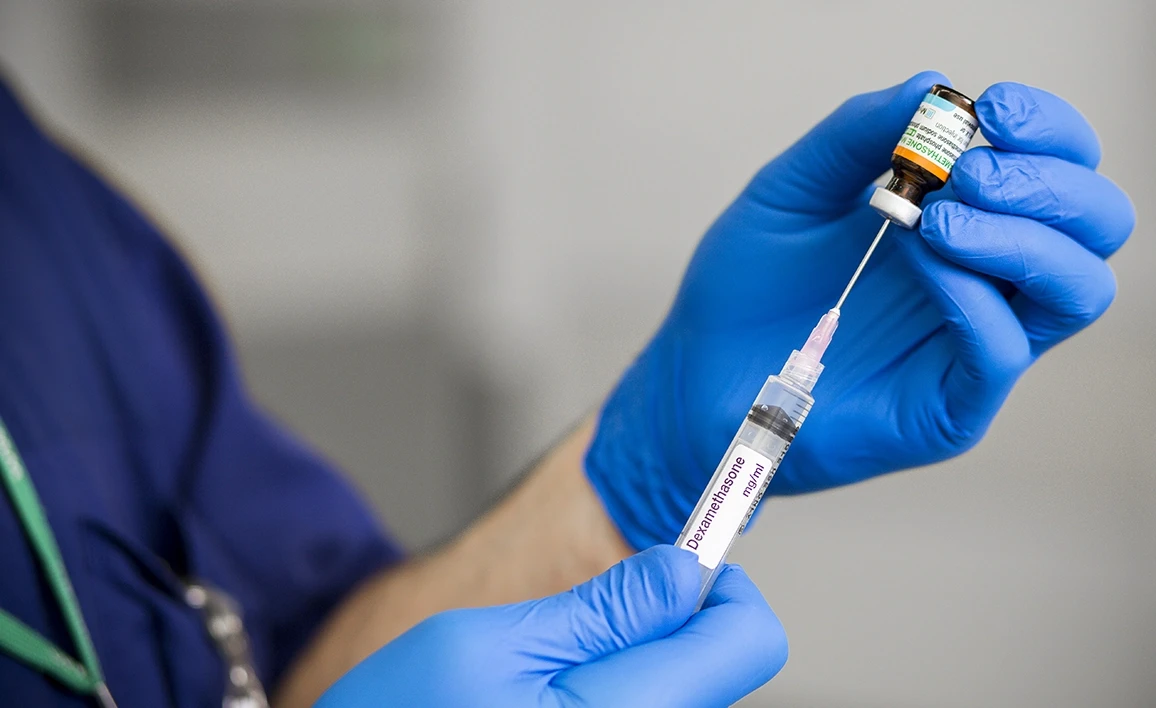 An anaesthetist in blue gloves prepares a syringe with Dexamethasone, drawing the medication from a vial in a clinical setting.