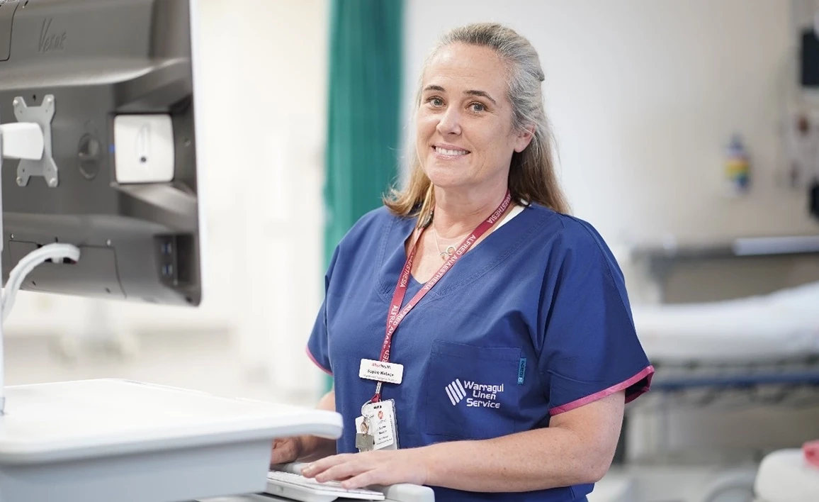 A smiling research coordinator in scrubs stands at a workstation in a hospital setting, ready to assist with patient care.