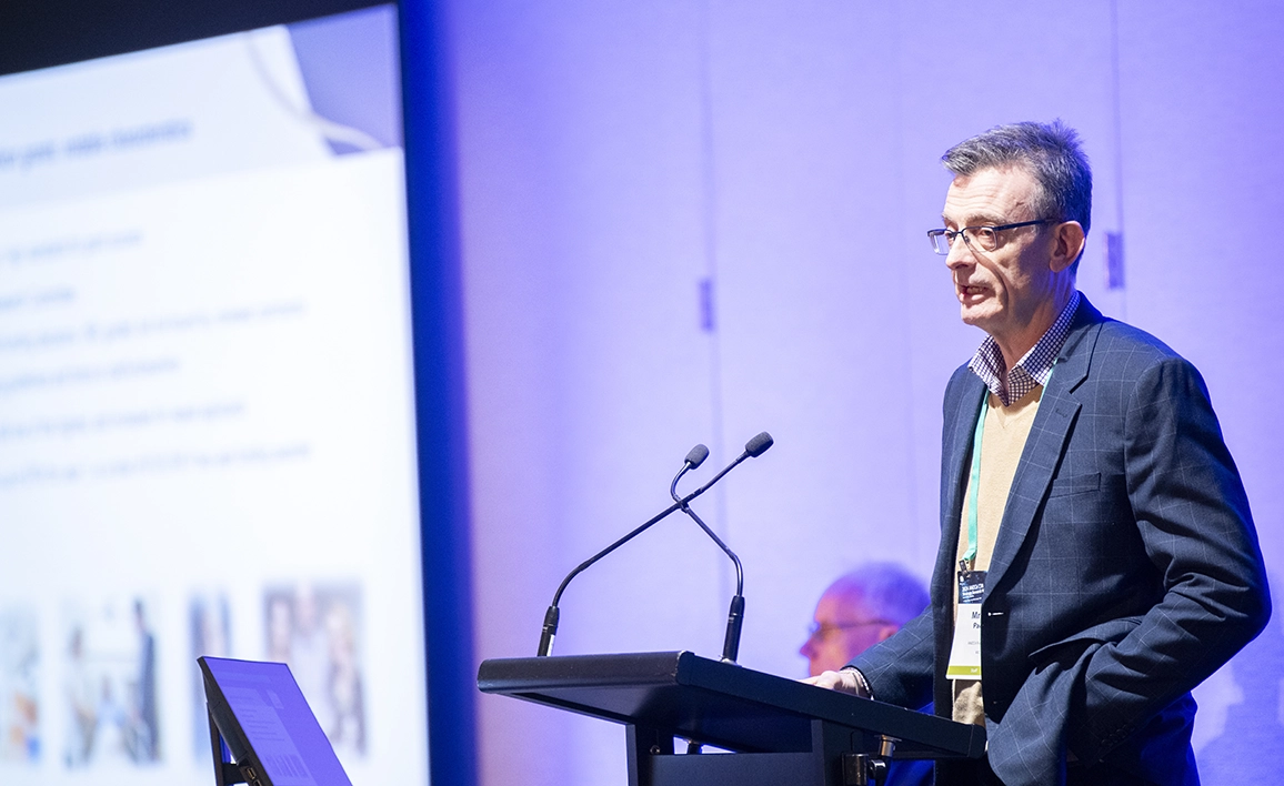 A professional speaker at a conference, presenting at a podium with a large screen displaying slides in the background.