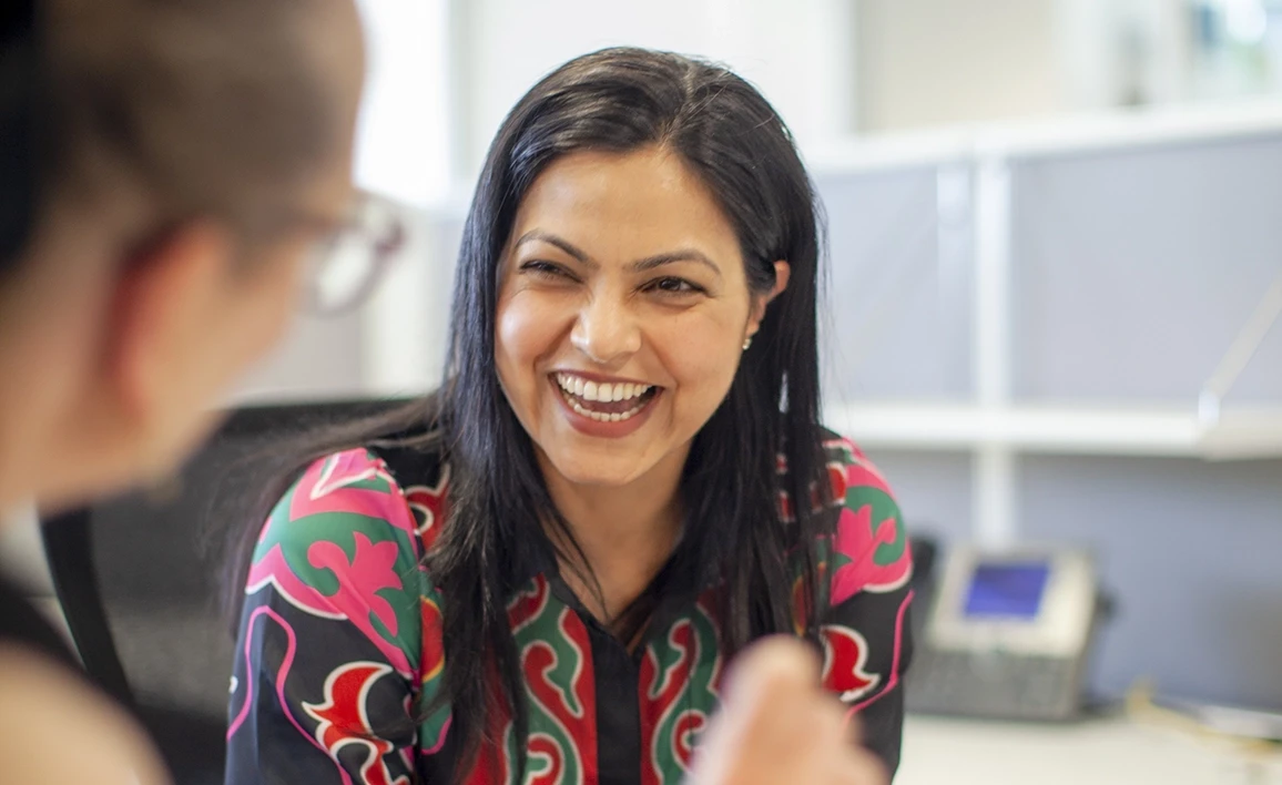 A smiling research coordinator in a colorful outfit engages in a conversation with a colleague in a bright, modern office setting