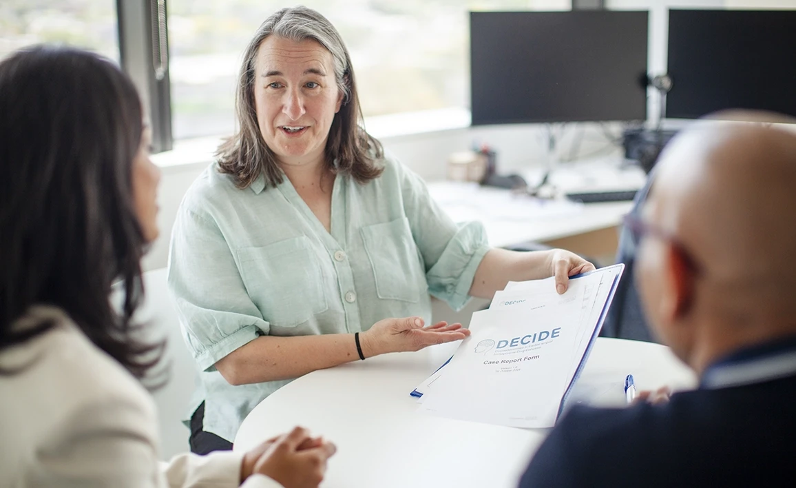 A research coordinator in a light green blouse discusses a document labeled "DECIDE Case Report Form" with a father and daughter in a modern office setting.