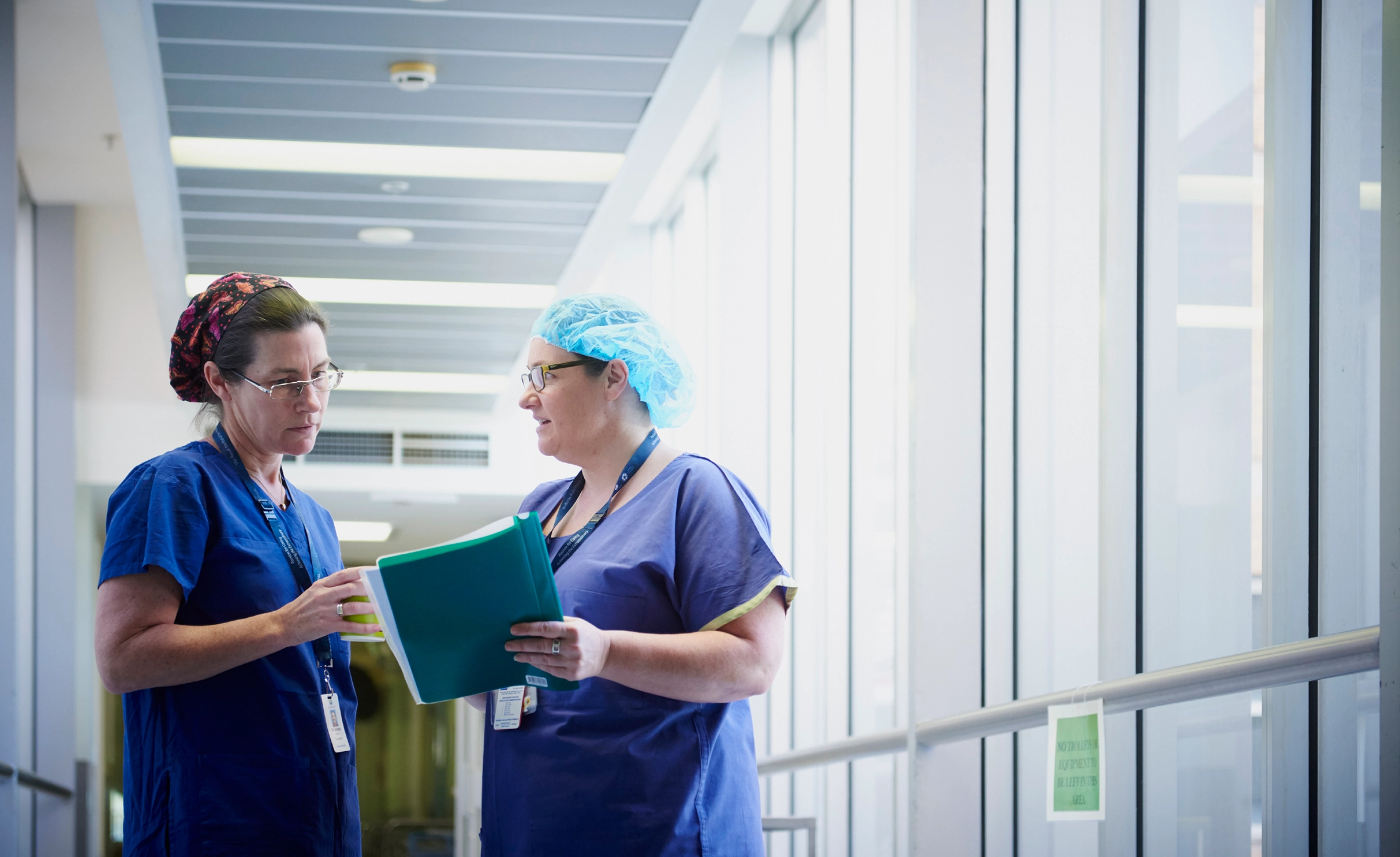 Two people in blue scrubs talking in a hospital corridor. One is holding a folder of papers