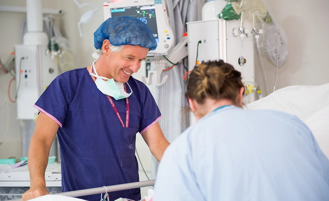 A smiling anaesthetist in scrubs and a surgical cap interacts with a colleague at a patient’s bedside in a hospital intensive care unit.