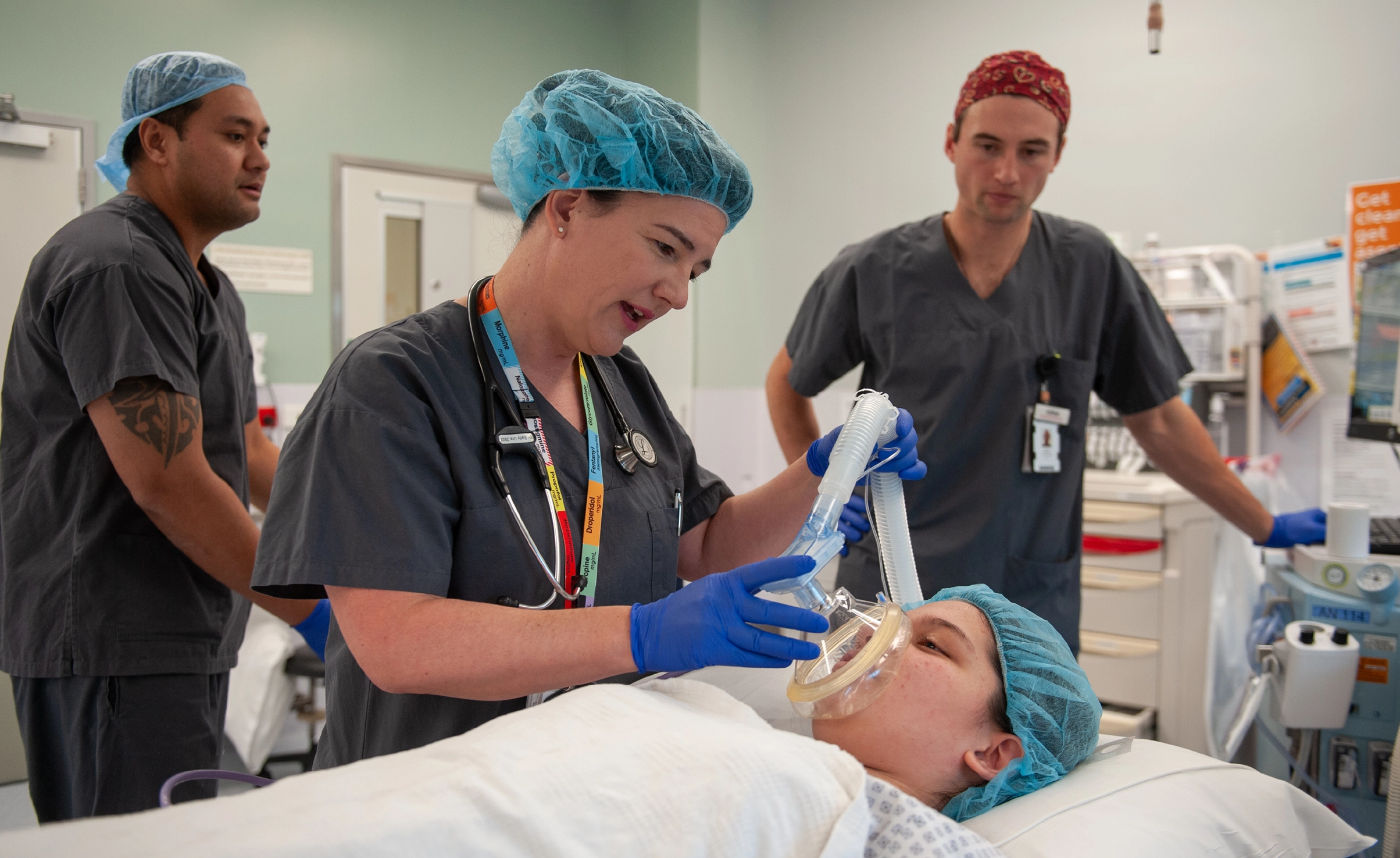 Anaesthetist in scrubs talking to patient in bed while giving anaesthesia via a mask while two people in scrubs look on in background