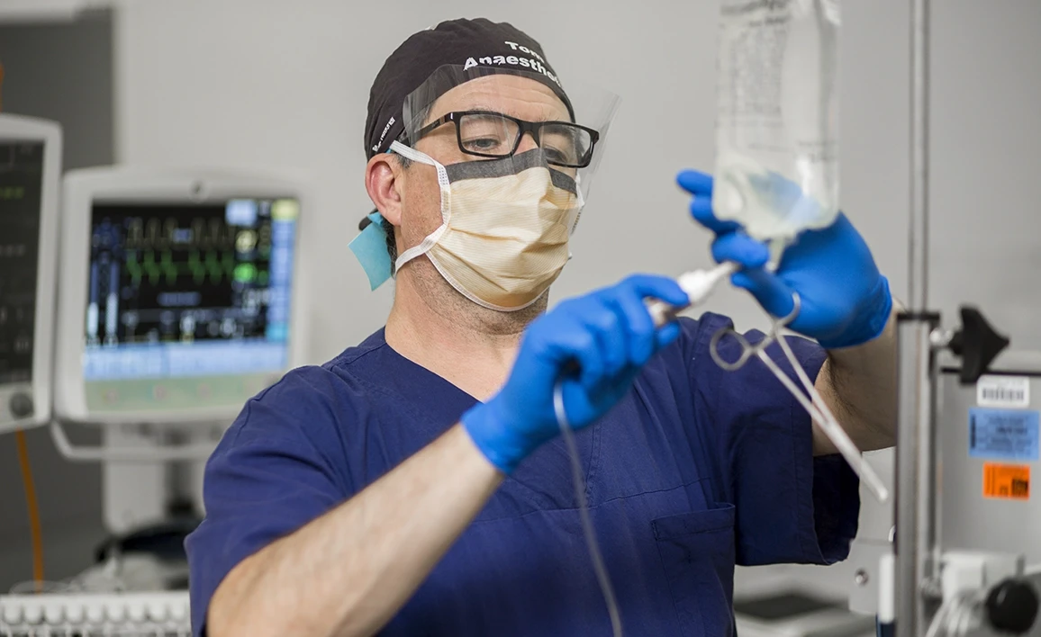 An anaesthetist in PPE prepares an IV infusion in an operating room, with medical monitors displaying vital signs in the background.