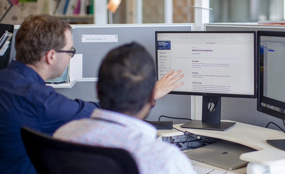 Two professionals at a workstation, discussing research information displayed on a computer screen in an office setting.