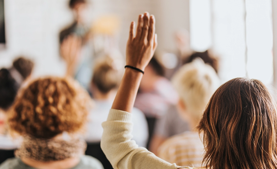 Woman raising hand in a crowd, facing a speaker